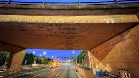 A concrete bridge over the M42 motorway. It has obvious wear and tear and marks on it. The motorway is closed with only highway vehicles parked up with their lights on. 