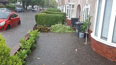 A residential street during severe flooding. Rain is falling, and floodwater covers both the road and the front garden area of a house. The water is deep enough to reach well above the kerb and has spread across driveways and pathways.