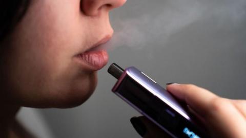 A woman holds a purple and blue vape near her mouth in a close-up image that only shows her hand and her lips.