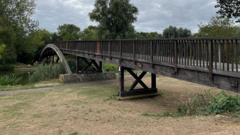 A wooden bridge stretches over a grassy area and a green river to another grass area on the other side.