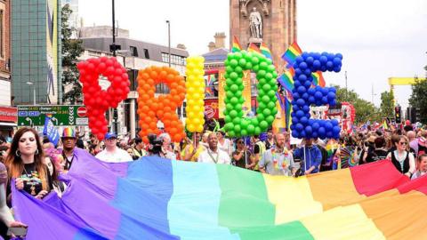 Large balloon letters that say Pride in rainbow colours held above a large rainbow flag.