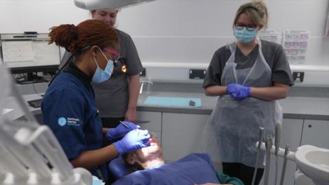 A person wearing a mask with their hair tied back has gloves on and is performing dentistry on a patient. The patient is lying down. There are two people watching over the procedure wearing clinical outfits.
