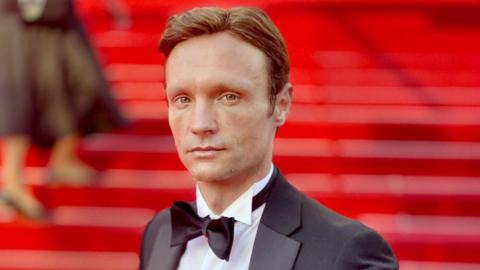 Hans Obma stood on a red carpet. Red stairs can be seen behind him, he wears a black suit jacket, white shirt and black bow tie. He has blue eyes and brown hair and looks side on down the lens. 