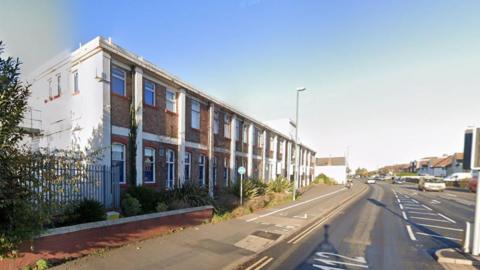 A white building with red bricks, and red wood around the windows. Opposite it is a road.