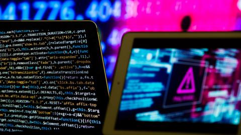 Composite image meant to represent computer software and hardware. On the left is a screen showing code in different colours. There is a pink hazard sign on the right of the picture.