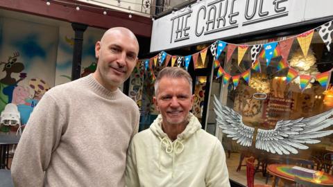 A man with a shaved head and grey jumper next to a smaller man with a grey quiff hairstyle and wearing a lemon-coloured hooded top.
