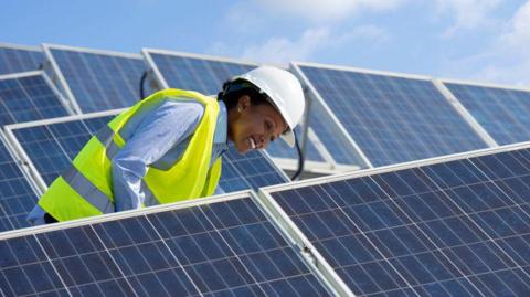 A woman engineer is checking solar photovoltaic panels at a solar farm. She is wearing a high-vis jacket and helmet and smiling.