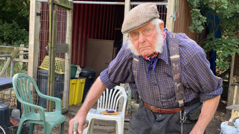 An elderly man wearing a flat cap, a shirt, a neck scarf and braces. He is standing using a walking stick. He is wearing glasses while looking directly into the camera.