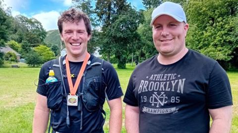 Two men smile side by side in a field. The man on the left has brown hair and wears a runners black vest with multiple pockets, a drink bottle tucked into his right breast pocket, and a black t-shirt. A square medal on an orange ribbon is around his neck. On the right is a man in a grey cap wearing a black t-shirt with white graphic print.
