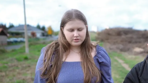 Image of Julia Wandelt walking through a field with her eyes down. She is wearing a purple dress and has mousey brown hair. 