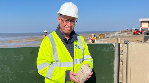 Dave Robson stands on Hunstanton pier. He is wearing a white hard hat and yellow high vis jacket. He is holding a core of concrete. 