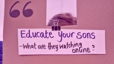A cardboard sign is held up to the camera, in marker pen it says 'Educate your sons - what are they watching online?'