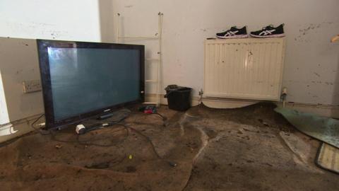 A flat screen TV on the floor on the left. The carpets are wet and are being stripped. There is a mop bucket at the back of the room next to a white radiator. 