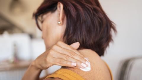 A woman applies a patch to her skin. She is wearing a yellow vest and she has straight brown hair. She is facing away from the camera.