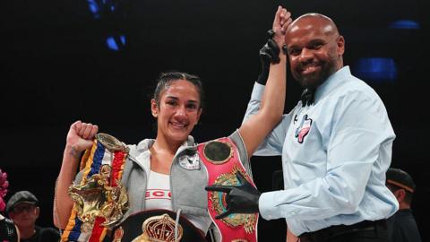 Amanda Serrano has her arm raised by a referee and is celebrating with her belt