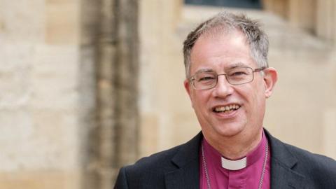 Bishop of Oxford, Dr Steven Croft is wearing a purple shirt, white dog collar and dark blazer. He has short greying hair and is wearing metal-framed glasses and is smiling towards the camera. The background is an out of focus wall. 