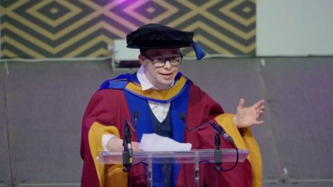 A man stands at a lecture wearing red blue and yellow robes. He wears black rimmed glasses and has a mortar board on his head.