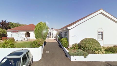 Longfield Villa is printed on the wall of a single storey building set on a  drive which leads to two other single storey buildings, one to the left and one behind. In the garden there are many plastic chairs set around garden tables and sun shades. There is a car parked off the road.