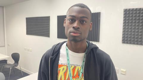 Tobi has very short black hair and black facial stubble. He is wearing a white t-shirt with a graphic comic book type design on it. He has a light blue lanyard around his neck and is wearing a dark navy zip hoodie