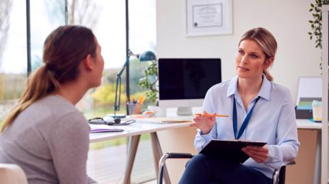 A female doctor in an office meeting with a female patient. They are sat opposite each other. The doctor has a clipboard in her lap and is holding a pen. Behind the pair are large windows that look out onto a garden.