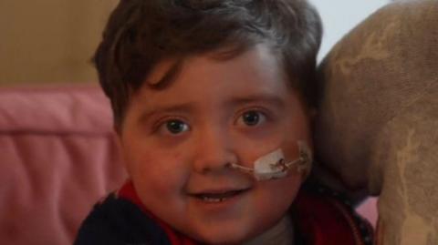 Ralph, who has short brown hair and has a nasogastric tube, smiles at the camera as he sits on the sofa next to his mother