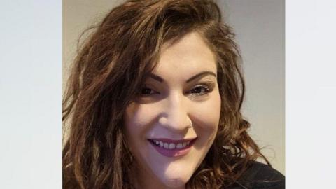 Portrait image of Charlotte Smith looking towards the camera smiling. She has dark brown hair in a side parting, and is wearing purple lipstick and a black T-shirt.