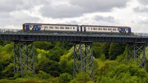 A Northern train running on the Northumberland Line crosses the North Seaton Viaduct. The blue and white two-carriage train cross over the grey steel bridge. It is passing over a large number of green, leafy trees.
