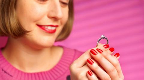 A woman on a pink background smiles while looking at a diamond ring.