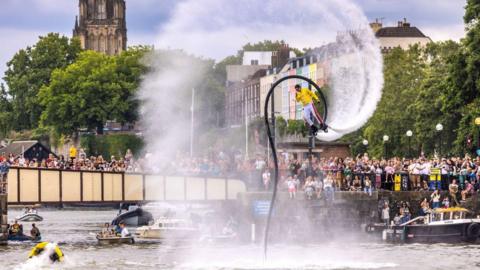 Image of a performer with a water jet pack suspended over the water in a harbour. There are a number of small boats in the water with people watching him. In the background is a bridge with a large crowd of spectators and the harbourside is also lined with spectators.