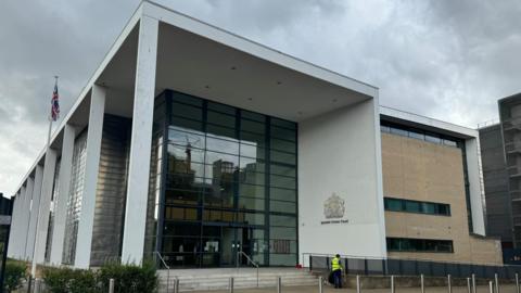 The front of Ipswich Crown Court. A large glass-panelled building with a white white pillars running down one side. It has a large silver sign that reads "Ipswish Crown Court" to its right.  