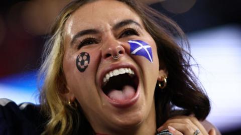 A female Scotland fan cheering. She has the flag of Scotland painted on her face