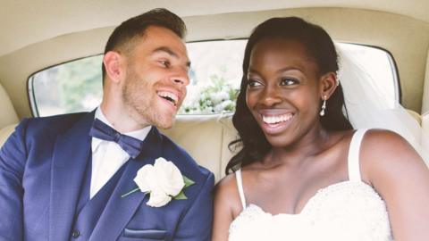 A man and woman in wedding attire in the back of a cream interior of a vehicle.