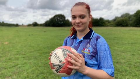 Isabelle standing in a field holing a red and white coloured football. She is wearing a blue Tottenham Hotspur shirt and has red hair.