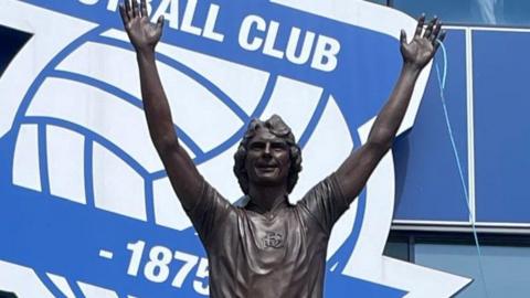 A bronze statue of Trevor Francis. The statue shows the footballer standing on one leg as if he is running, with his arms in the air, as if he is celebrating a win. The stadium's football club sign is in the background. There are people standing around the sculpture as it is unveiled.