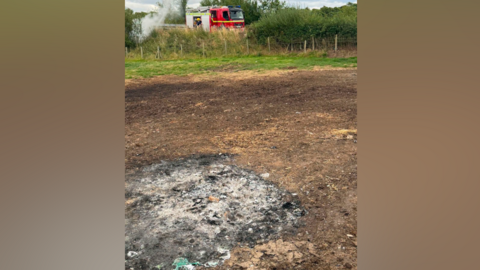 A patch of land which has been scorched and there is a black patch of ashes. It is on countryside land and in the distance there is a Cumbria Fire and Rescue appliance which a firefighter is exiting from.