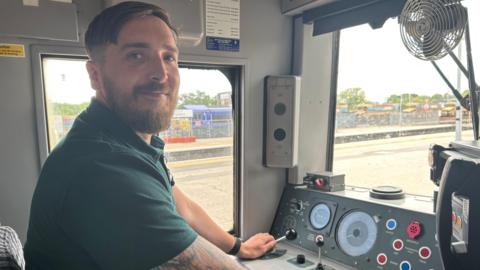 Ben Warner smiles at the camera while sitting in a drivers cab for a train. Lots of dials and leavers can be seen. He wears a green polo shirt.
