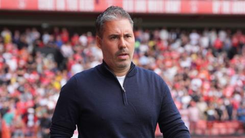 Marti Cifuentes looks ahead with a determined and assured expression on his face. He wears a navy quarter zip top and there's a crowd of people inside a stadium in the background.