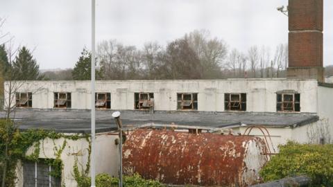 A derelict white building with a large, rusted metal container in front of it. To the right is a large red brick chimney