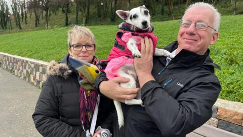 Paul is holding Jessie the Jack Russell Terrier in his arms up on his chest as he stands in a park. He is wearing a black coat and glasses. He has grey slicked back hair. Jessie is wearing a pink dog coat. Lola is standing next to Paul and she has a dark coat on with a fur lining the hood. She has a multi-coloured scarf and has short hair and glasses.