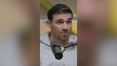 A man with slicked back dark hair and wearing a light grey hoodie speaks into a microphone.