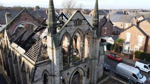 An aerial view of Park Church. The building has no roof. Rubble and plant life can be seen inside the building. Three large windows stand empty where there used to be stained glass. The building has been built in the Gothic Revival style.