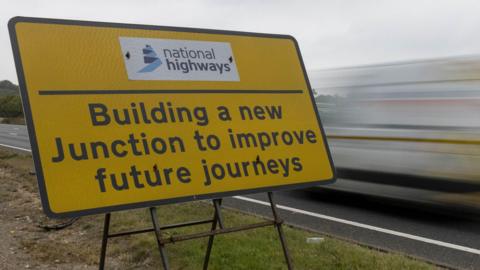Close up of a yellow sign on a grass verge with the words BUILDING A NEW JUNCTION TO IMPROVE FUTURE JOURNEY written in black. A blurred vehicle drives past the sign on the right