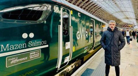 A man with grey hair is wearing a coat on the right hand side of the photo. The image is dominated by a green train at a railway station. On the left of the image the train has the words Mazen Salmou on it.