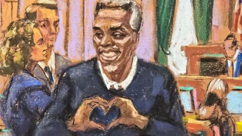 A court sketch of Diddy smiling, making a heart shape out of his hands as he stands in the courtroom