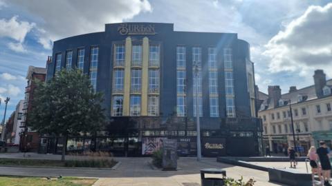 A large dark-coloured multi-storey building with 25 visible windows and a sign that reads "Burton". There are trees and other buildings in the foreground with paving and people walking by.