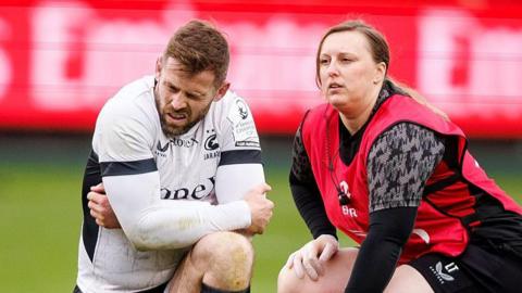 Elliot Daly (left) kneels on the pitch with his arms bent across his knee clutching his left elbow, with a grimace on his face during Saracens' defeat by Bath on 4 April