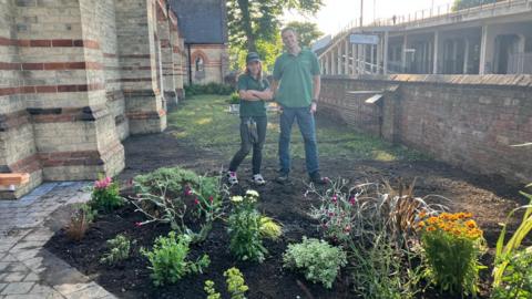 Woman wearing green T-shirt, cap and dark jeans with man wearing green T-shirt with blue jeans stand on a patch of soil behind plants, next to a paved area. There is a brick and stone church building to one side of them and a brick wall and another building to the other.