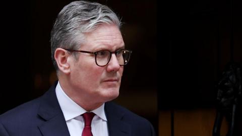 British Prime Minister Keir Starmer wearing a suit and red tie looks sternly out of the door to 10 Downing Street