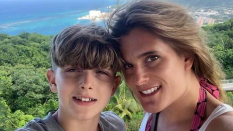 Julian Sweeney and his mother, Ellen Roome. They both have light brown hair and are pictured overlooking a view of trees and sea. Both of them are looking at the camera and smiling.