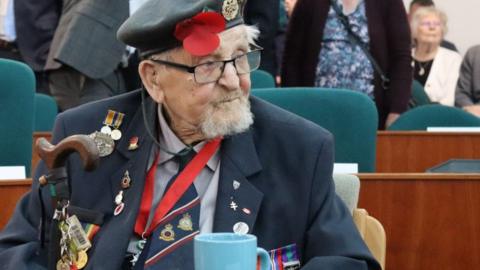 An elderly male war veteran sits in a council chamber with his uniform, including a navy beret, blazer and navy, red and white striped tie, adorned in badges and medals. He has a walking stick and is wearing a red lanyard and black glasses. He has a blue mug in front of him.
